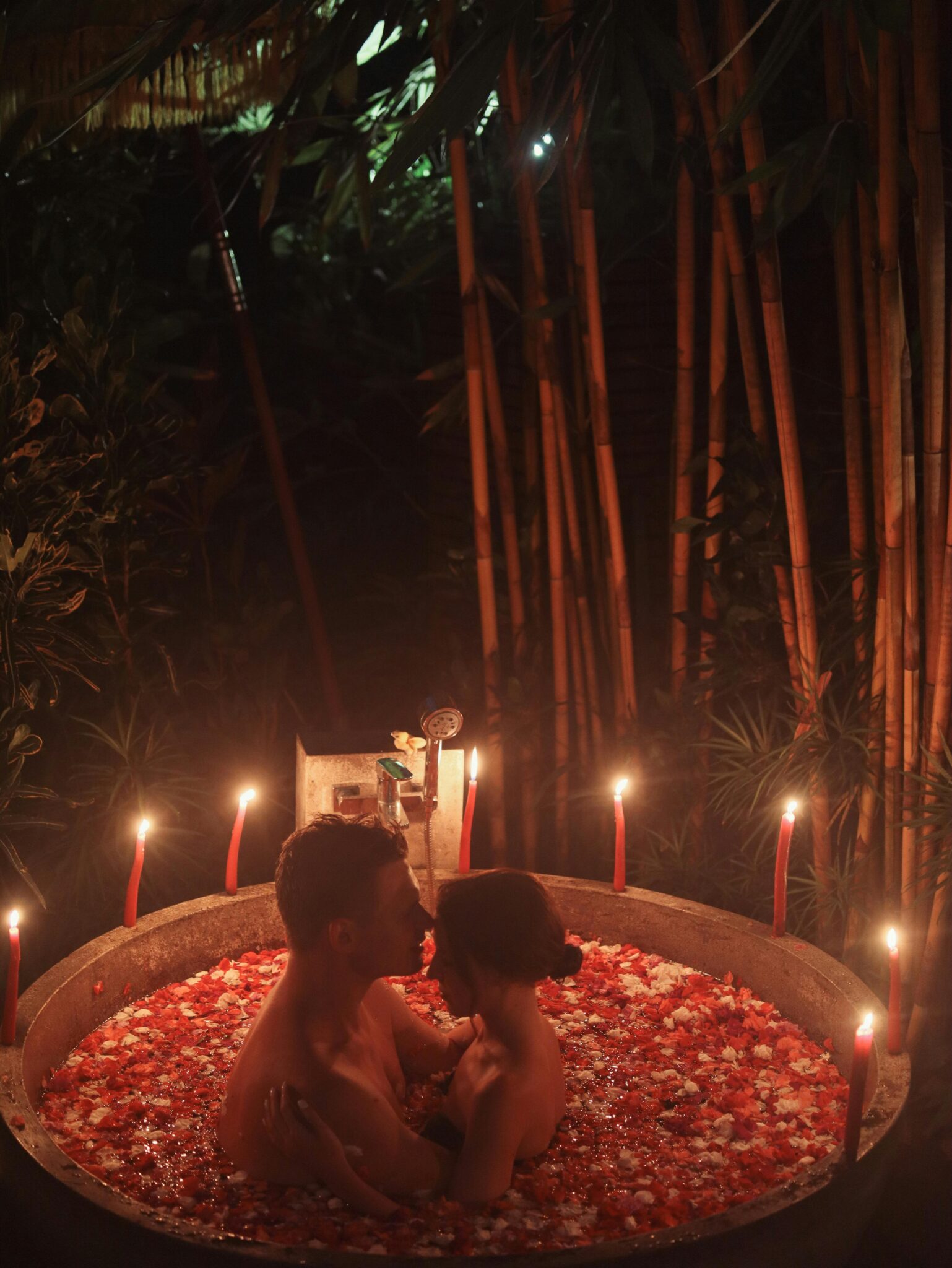 From above of sensual couple in romantic atmosphere hugging passionately and resting in round warm bath with rose petals surrounded with candles in tropical resort at night